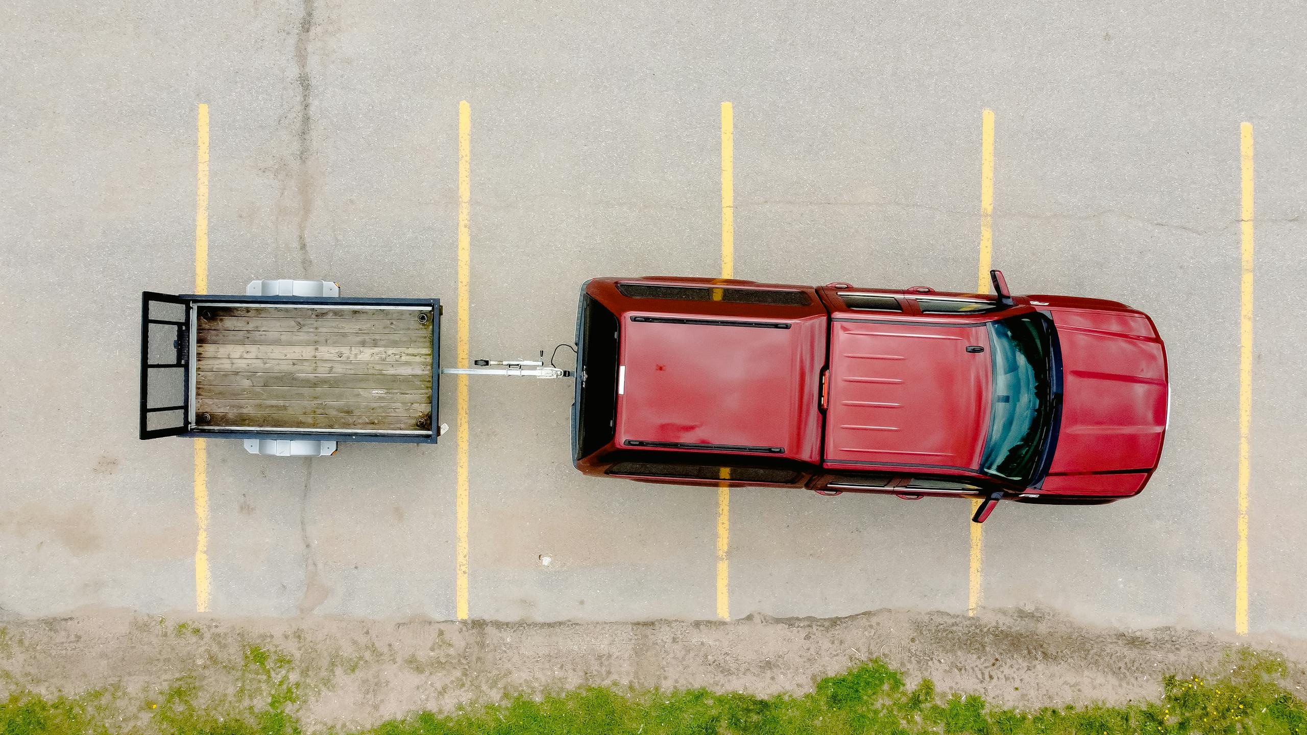 Aerial shot of a red car with a trailer parked on an empty lot with visible parking lines.