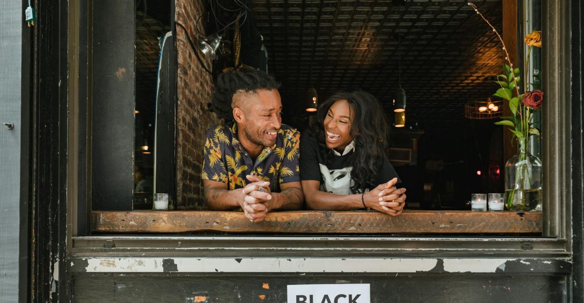 Two joyful business owners lean on a window sill with a Black Businesses Matter sign visible