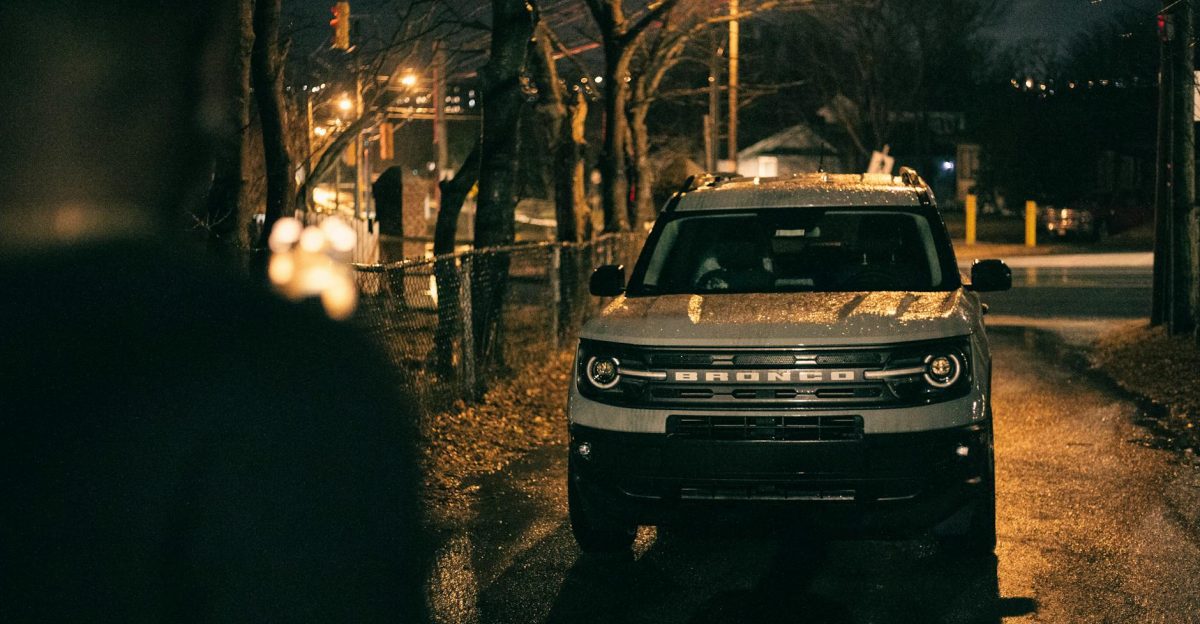 A white SUV parked on a wet urban street at night surrounded by dim streetlights and trees