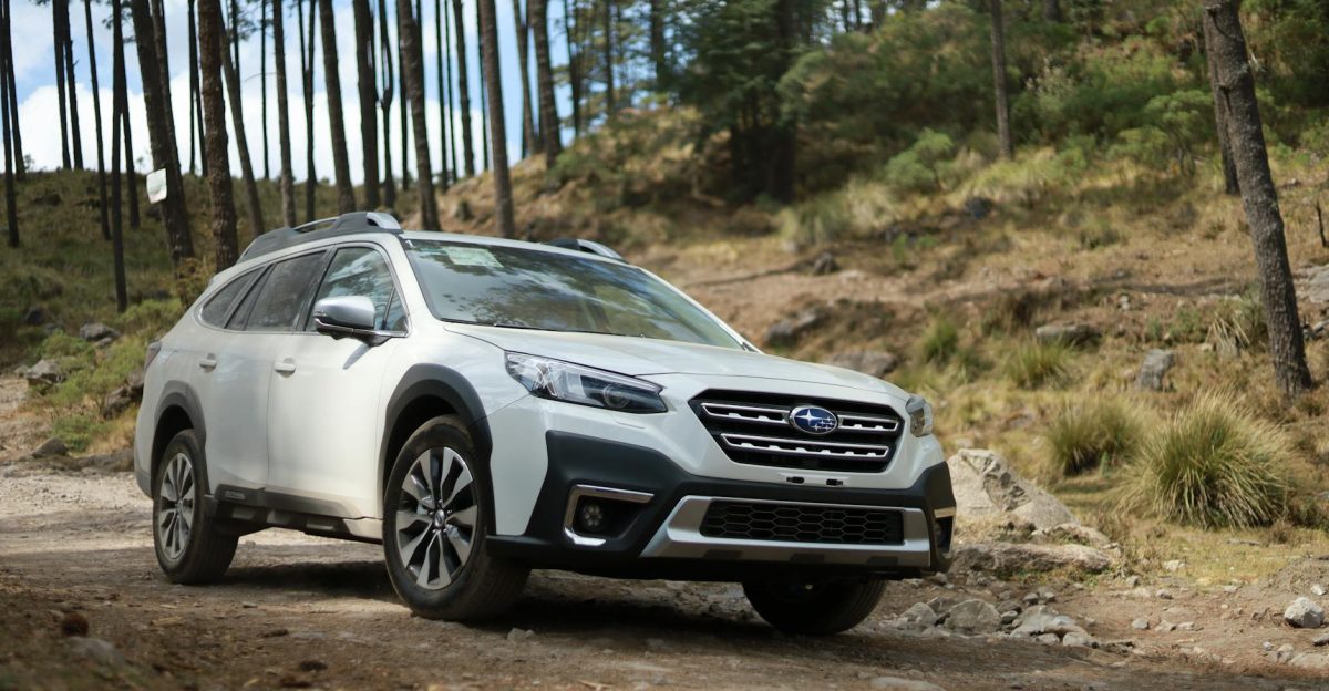 A white Subaru Outback SUV driving on a rugged forest trail among tall trees