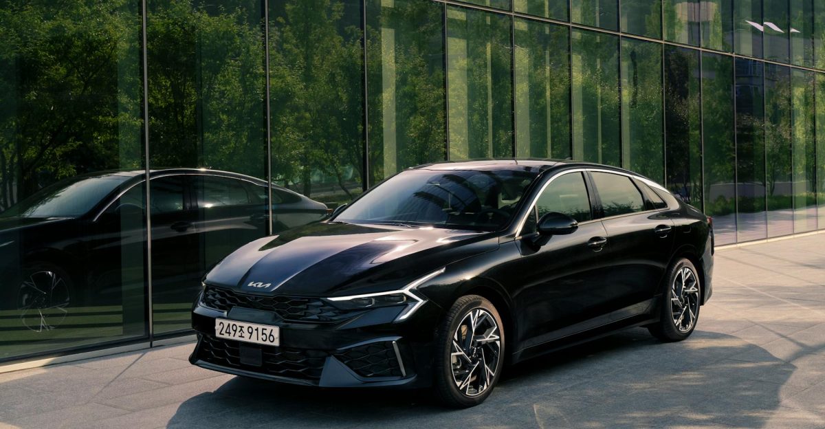 A stylish black Kia K5 sedan parked in a modern urban area reflecting city architecture