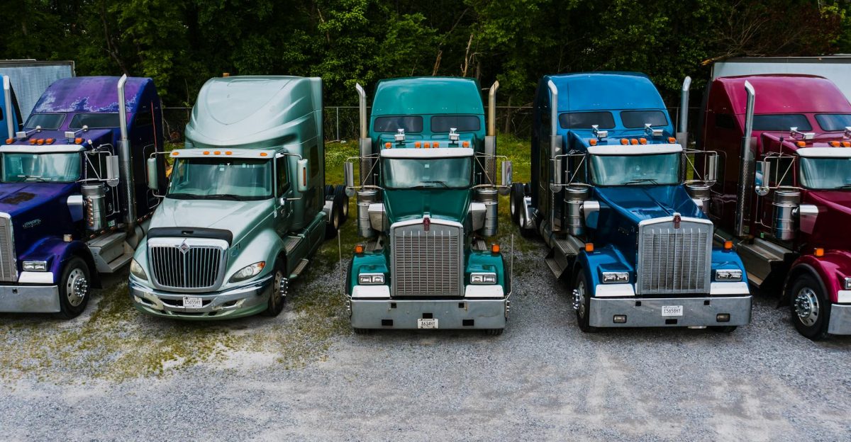 Row of vintage classic trucks of different colors parked on roadside outside warehouse against lush trees
