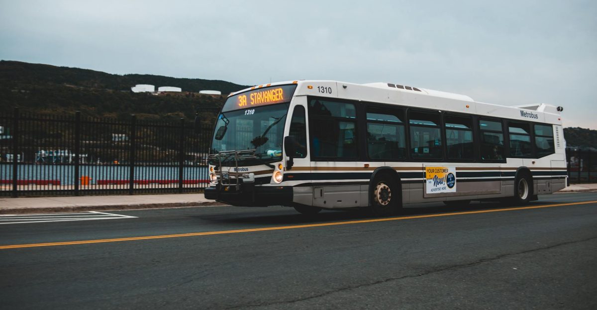 Big gray bus driving on asphalt highway along metal fence near water against cloudless sky with mountains in distance outside