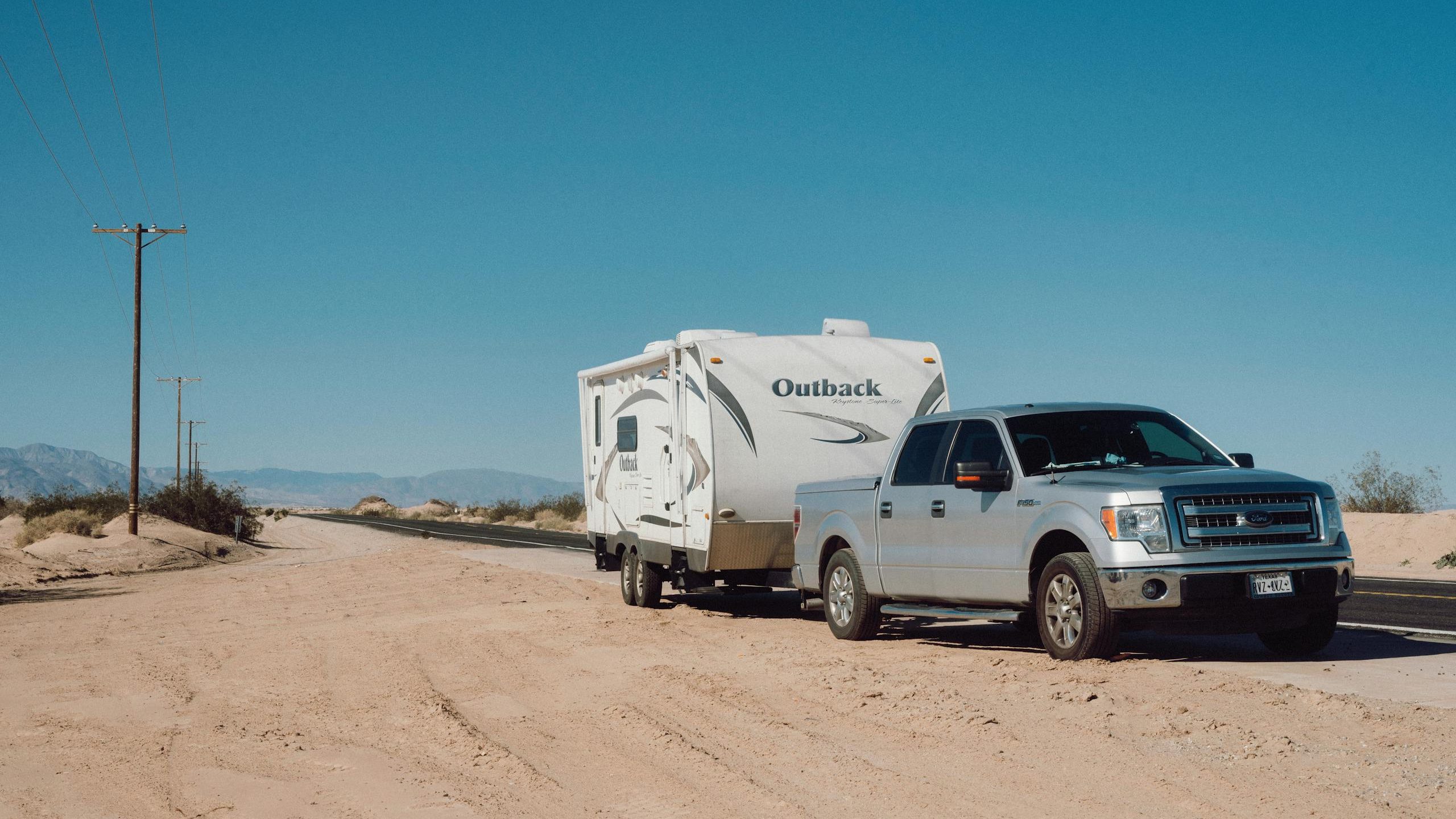 Silver truck towing an Outback trailer on a desert road under clear blue skies.