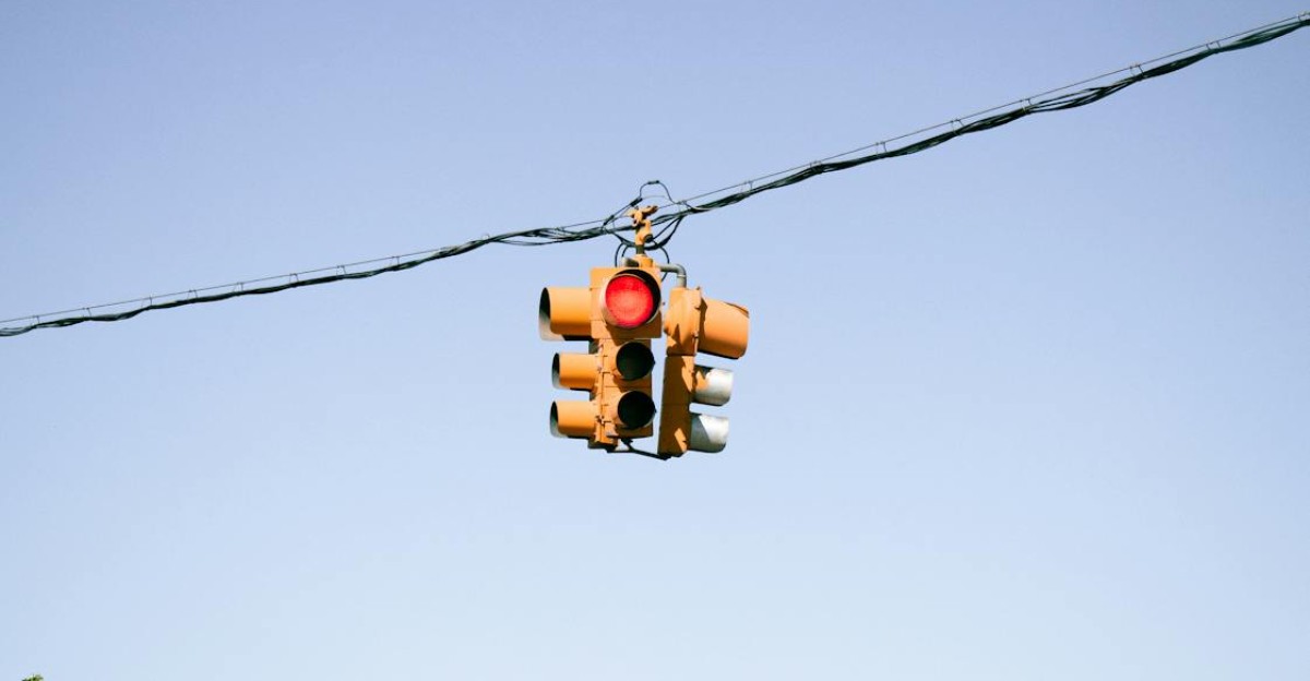 A traffic light with a red signal hanging against a clear blue sky overhead wires visible