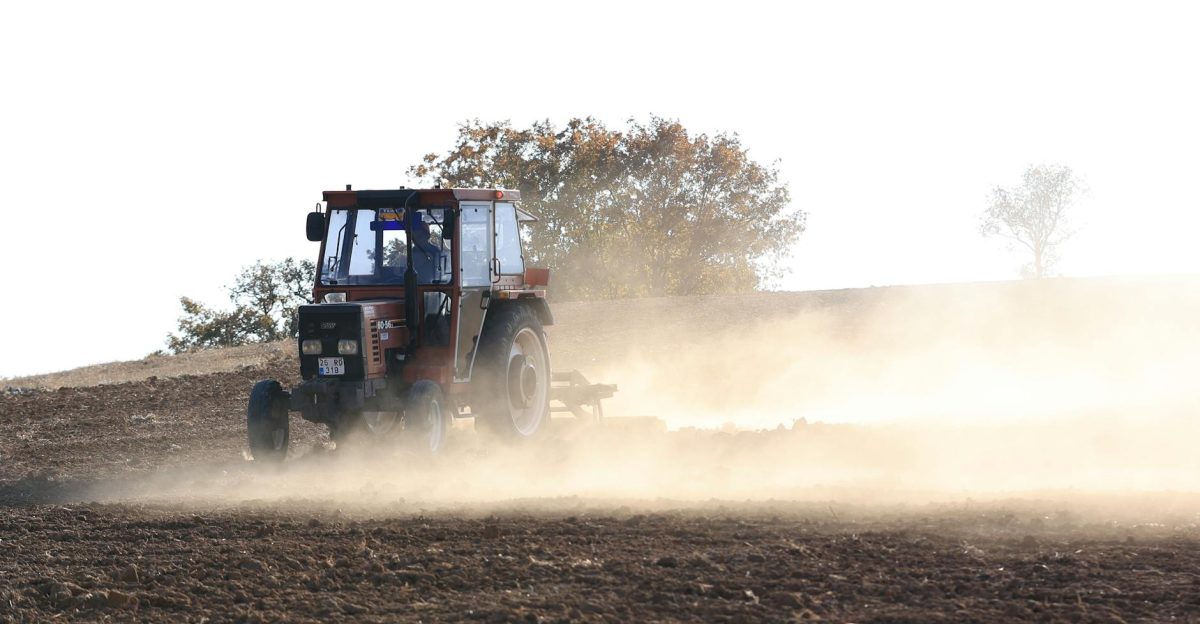 A farmer drives a red tractor through a dusty field preparing for planting