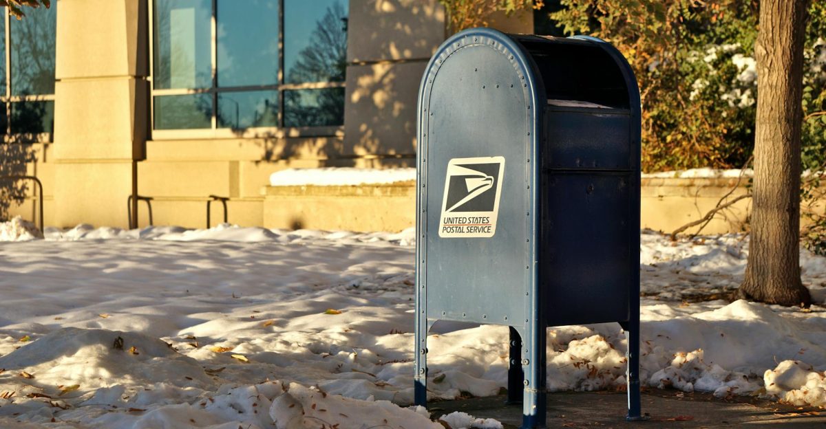 A USPS mailbox stands in a snowy urban area during winter sunlight casting shadows