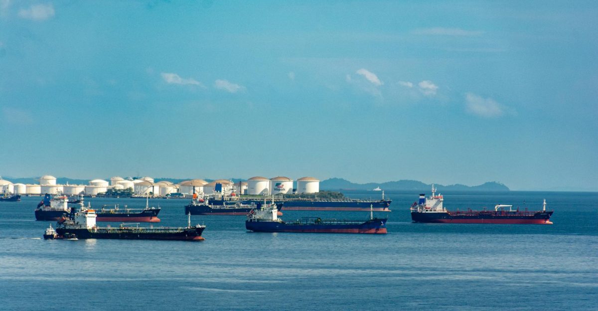 A fleet of cargo ships docked near oil storage tanks along a serene coastline with a clear blue sky above