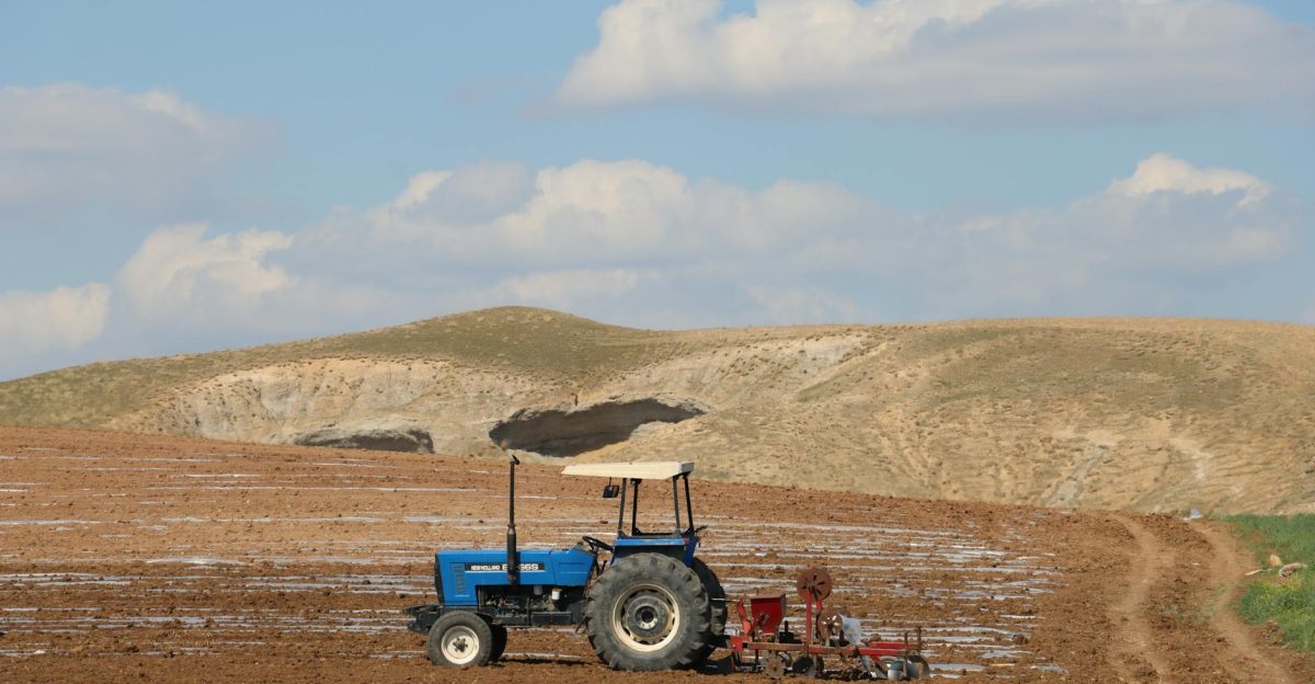 A blue tractor plowing a vast farmland against a backdrop of rolling hills and clouds