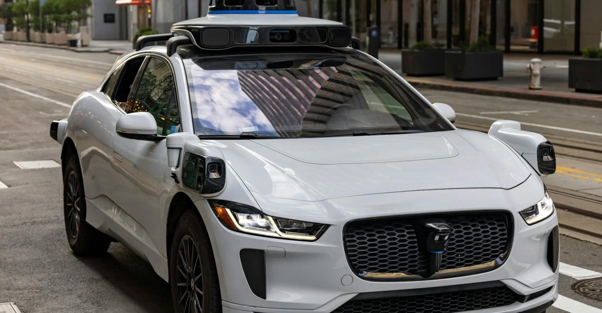 A white autonomous vehicle navigating a city street reflecting urban architecture in daylight