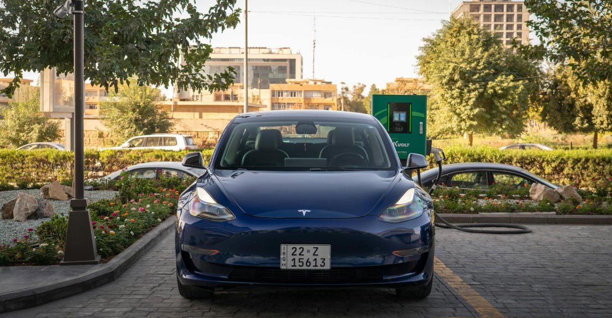 A blue Tesla Model 3 charges at an outdoor station during the day surrounded by greenery and urban architecture
