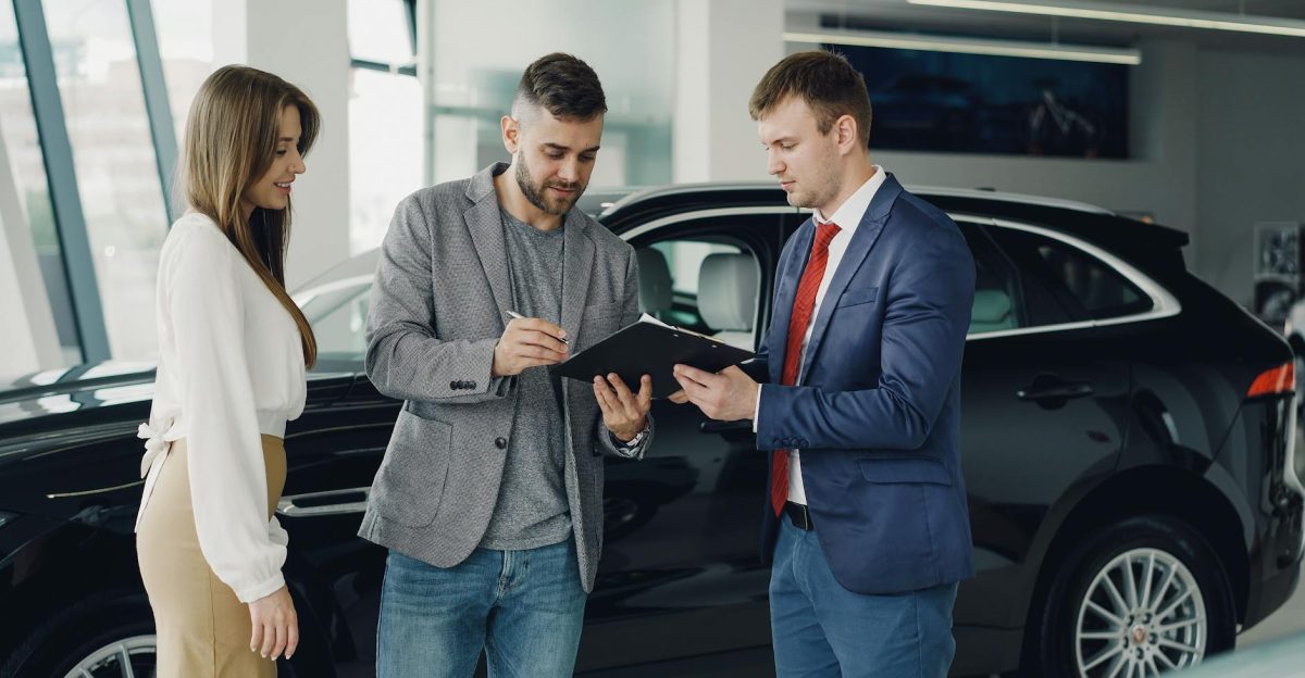 Three adults discussing documents at a car dealership beside a black car