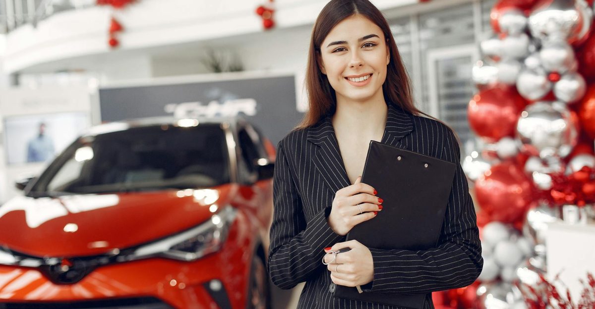 Cheerful friendly stylish female consultant in trendy black formal dress standing with clipboard standing in car showroom against new red shiny car and looking at camera