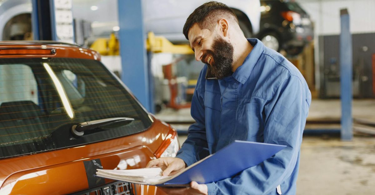 Smiling auto mechanic inspects a car in a service garage wearing coveralls and holding documents