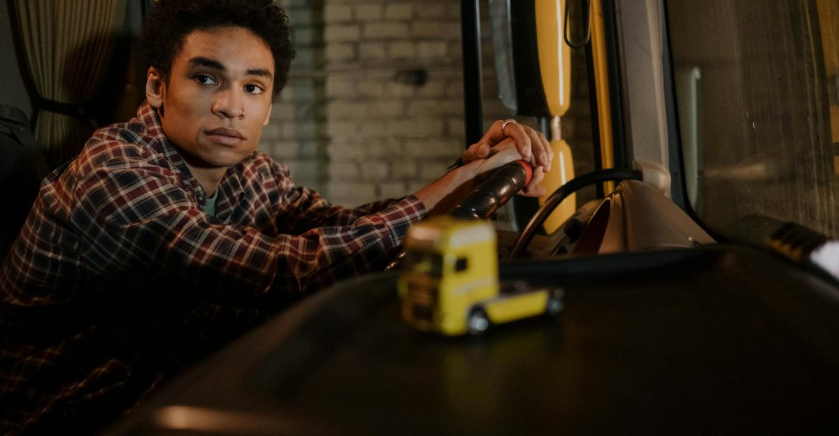 A young man sits in the cabin of a truck hands on the steering wheel with a model truck on the dashboard