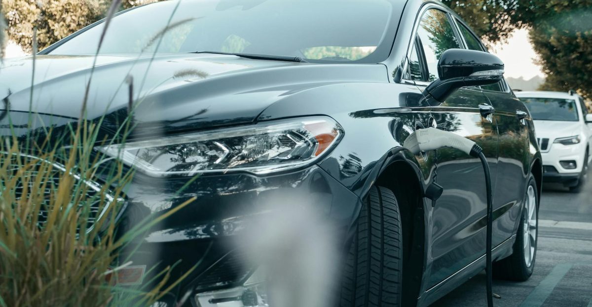 A black hybrid car charging at an outdoor station surrounded by greenery